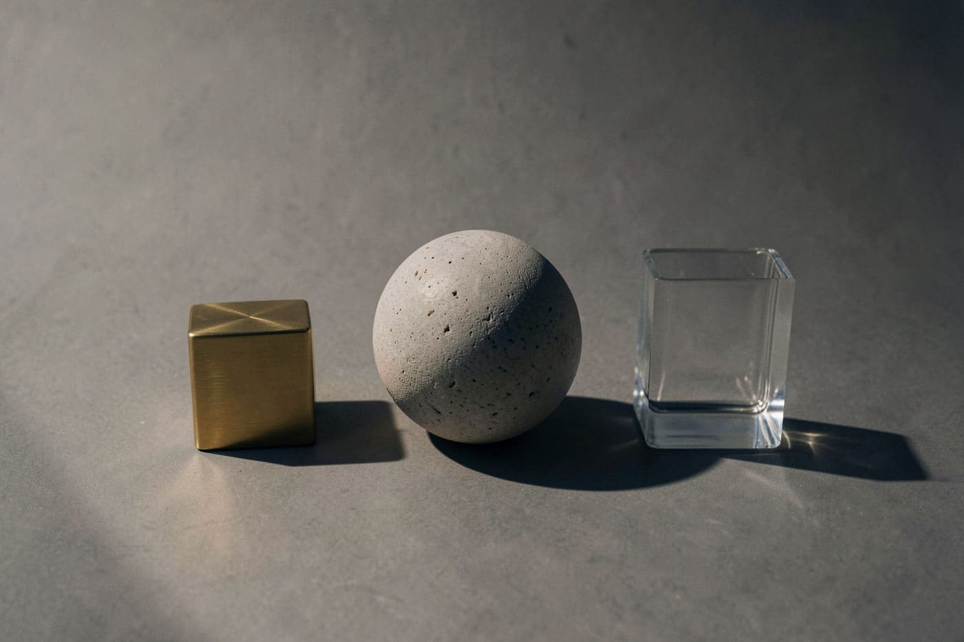 A brass cube, a limestone sphere and a glass block in a row under hard light.