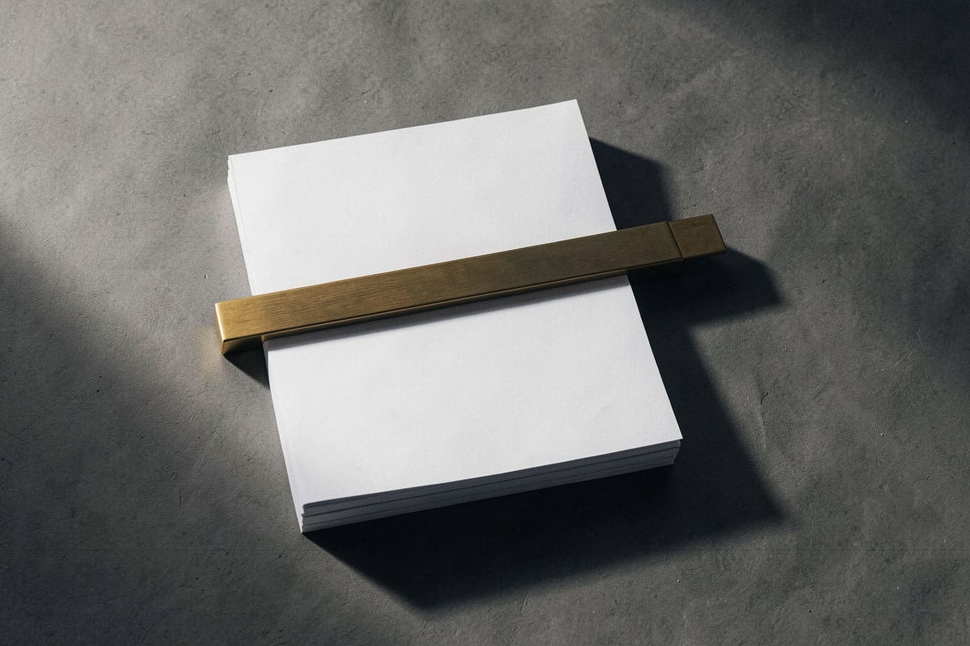 A thick stack of blank paper on warm stone-grey, with a single brushed brass bar laid across it as a weight.