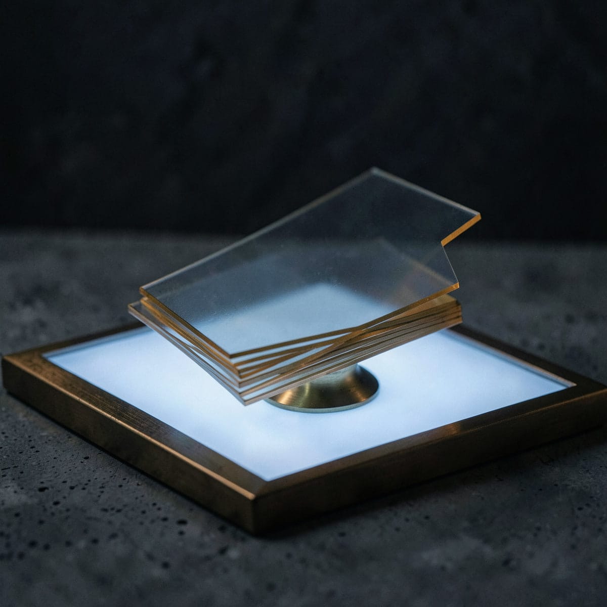 A fanned stack of thin translucent cards with brass edges on a glowing light table, one card lifted above the rest on a small brass stand.