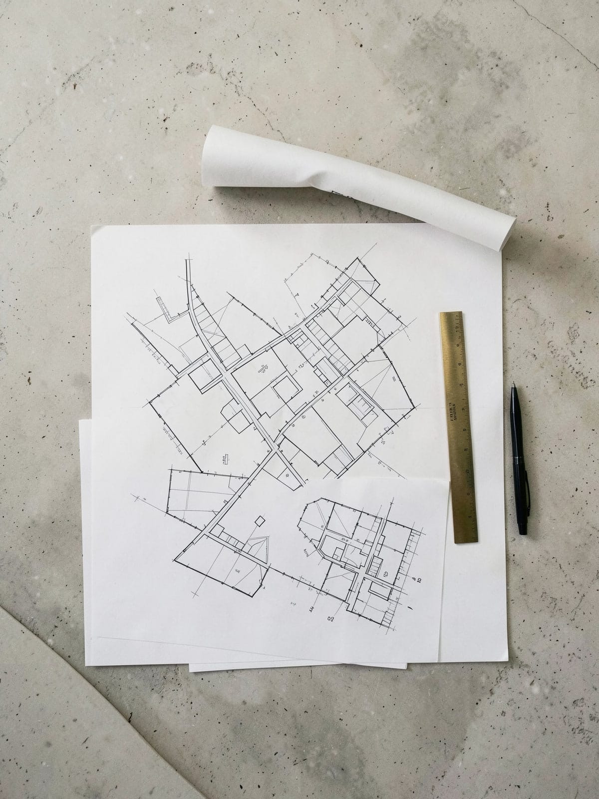 Architectural site and floor plans laid out on a limestone table, a rolled drawing above them and a brass scale and pen alongside.