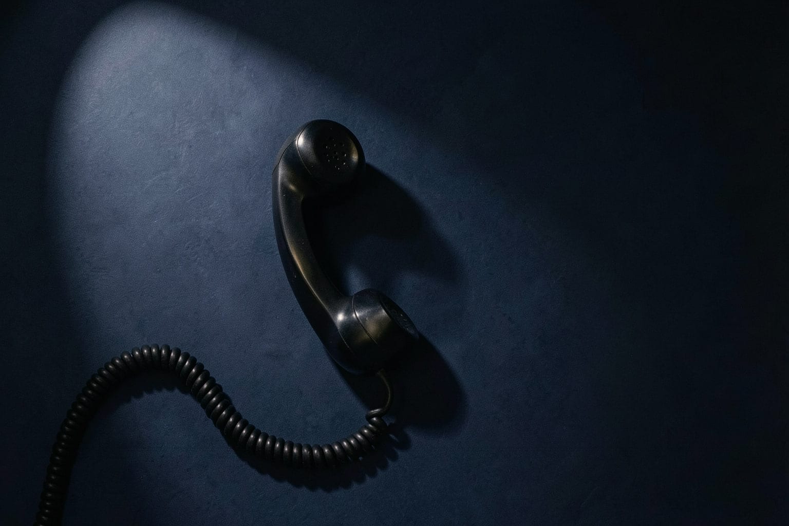 A black telephone receiver on its own, its coiled cord curling away under a single hard light.